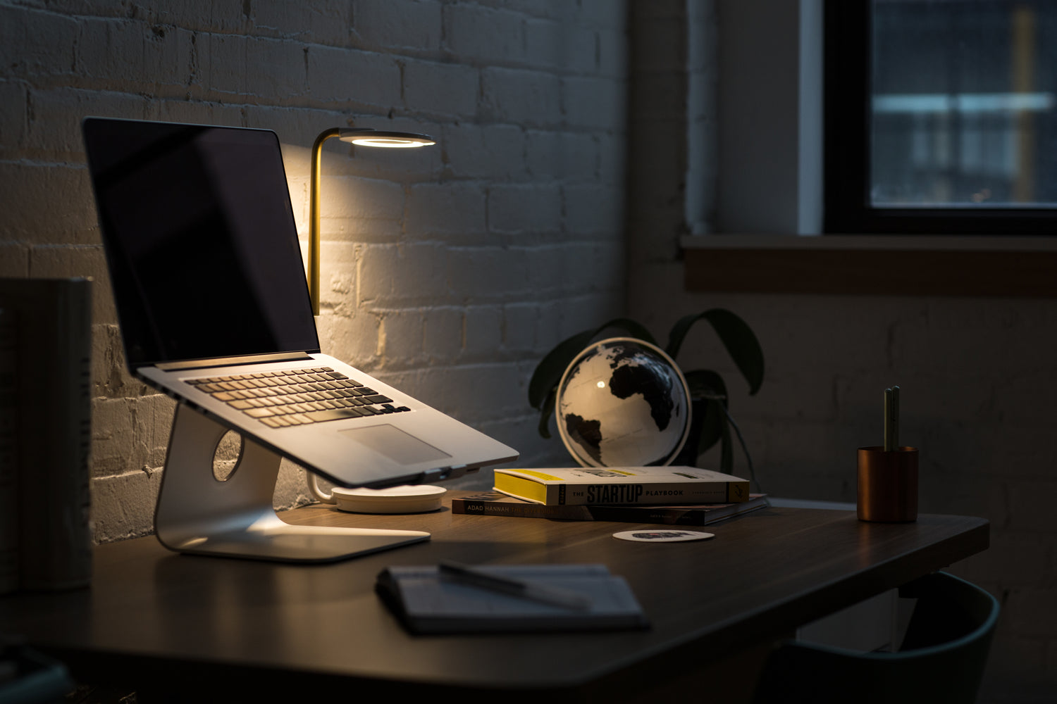 a laptop sits on a desk in a dim room with a desk lamp over it. There are some generic desktop objects on the desk too.